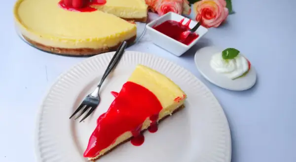 Ein Stück New York Cheesecake mit roter Fruchtsauce und Himbeeren, daneben eine kleine Schüssel Sauce und ein Klecks Sahne.