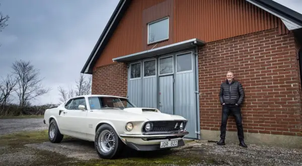 En man står stolt bredvid sin vita Ford Mustang Boss 429 framför en röd tegelbyggnad.