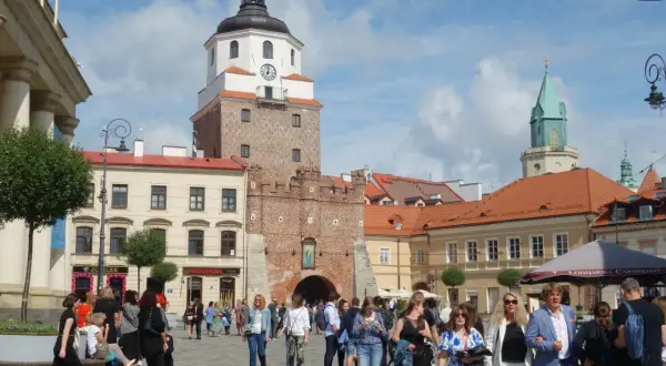 Ludzie spacerują po brukowanym placu litewskim, podziwiając zabytkową wieżę z zegarem i zieloną iglicę w tle.