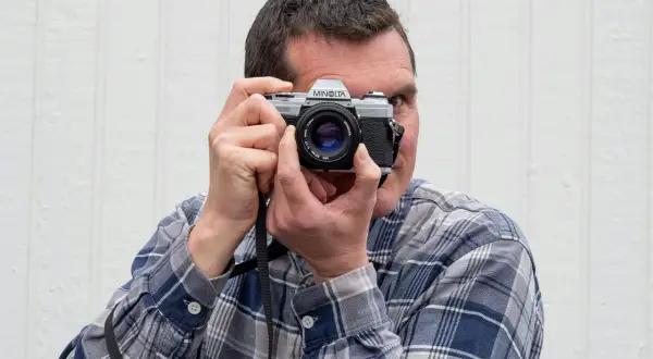 Man med rutig skjorta fotograferar med en Minolta-kamera, en motsats till analog teknik.