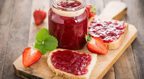 Marmellata di fragole fatta in casa con il Bimby, spalmata su fette di pane fresco, con fragole intere e a metà.