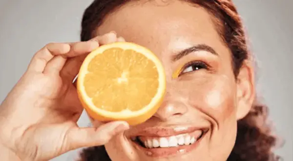 Strahlendes Lächeln mit einer Orangenscheibe vor dem Auge, die Frische und Vitamin C für die Hautpflege symbolisiert.