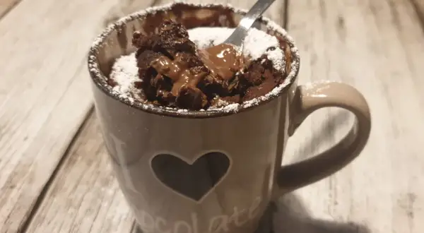 Un delizioso mug cake al cioccolato, appena sfornato, con cuore e scritta "chocolate" sulla tazza, spolverato di zucchero a velo.