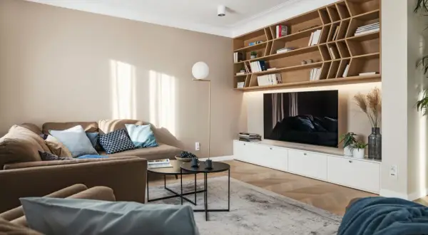 Przytulny salon z sofą i regałem, gdzie dominują kolory pasujące do beżu i brązu. Jasne ściany i drewniane elementy tworzą ciepłą atmosferę.