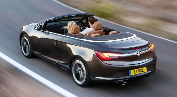 Para podróżuje lśniącym, ciemnobrązowym **Opel cabrio** po krętej drodze.