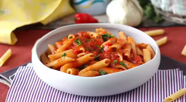 En skål med pasta arrabiata, toppad med färsk persilja. En vitlöksklyfta och en röd chili syns i bakgrunden.
