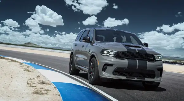 Szary Dodge Durango SRT Hellcat z czarnymi pasami pędzi po torze wyścigowym pod błękitnym niebem z chmurami.