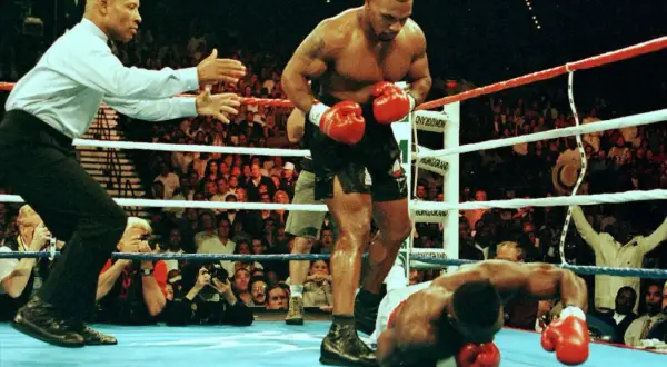 Mike Tyson stands over a fallen opponent, his powerful physique and impressive mike tyson arm length evident. The referee signals the end of the match.