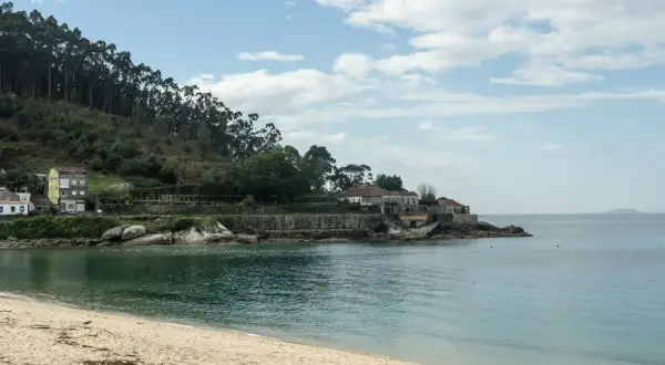 Arena dorada y mar turquesa en la playa de Loira, con casas blancas y una colina boscosa al fondo.