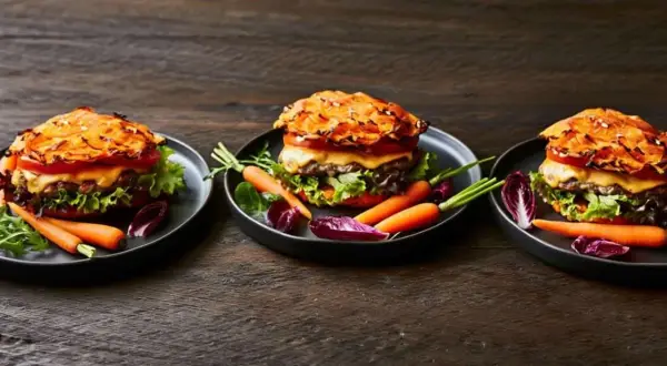 Drei appetitliche Hamburger mit Pommes, garniert mit Karotten und Radicchio auf dunklem Holztisch.