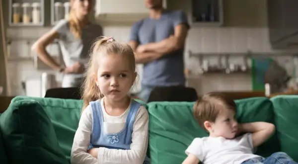 Zazdrość u dzieci w wieku szkolnym. Dziewczynka z założonymi rękami patrzy w bok, obok siedzi chłopiec. Rodzice w tle.
