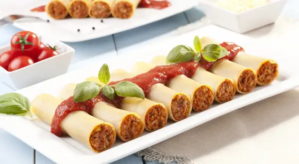 Cannelloni z mięsem i sosem pomidorowym, udekorowane bazylią, na białym talerzu.
