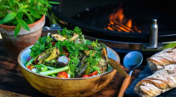 En härlig somrig sallad med grillade grönsaker, tomater och rädisor i en träskål. Bredvid ligger nybakat bröd och en grill med flammor i bakgrunden.