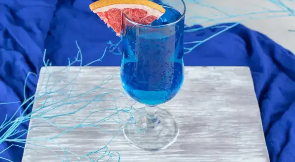 Niebieski drink, jak orzeźwiająca blue lagoon, z plasterkiem grejpfruta, na drewnianej desce, otoczony błękitnymi gałązkami.