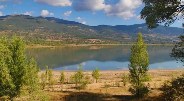 El embalse de Pinilla, con montañas verdes al fondo y un cielo azul con nubes blancas. Reflejos en el agua serena.