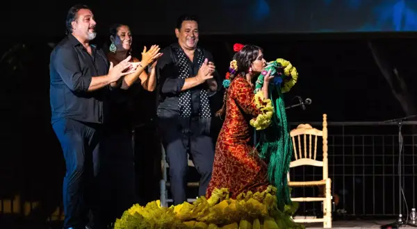 Una bailaora de flamenco andalusi, con un vestido rojo y amarillo, besa un mantón verde. A su lado, tres músicos aplauden.