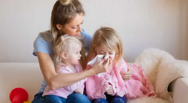 Mama troszczy się o córkę z katarem, podczas gdy druga córka patrzy. Czy szczepienie mogłoby temu zapobiec?