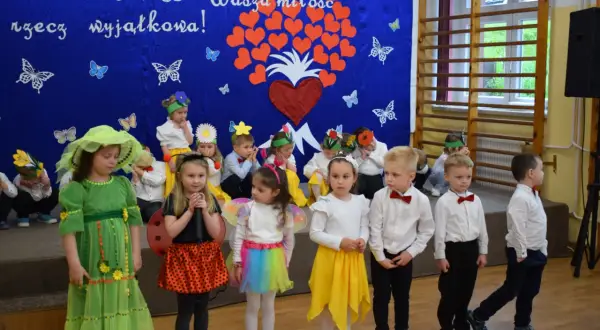 Przedszkolanka w zielonej sukience i dzieci w strojach na tle niebieskiej kurtyny z sercami i motylami.