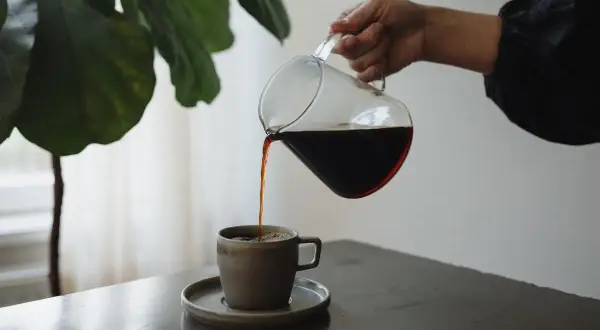 En hand häller upp mörkt kaffe ur en glaskanna i en mugg. Kaffet innehåller njutning och energi.