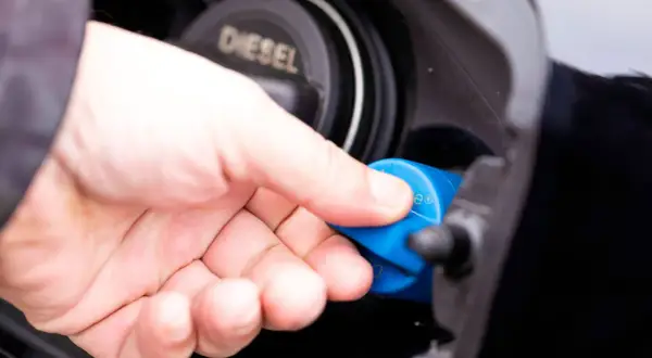 Hand schraubt blauen AdBlue-Deckel ab. Diesel-Tankdeckel daneben.