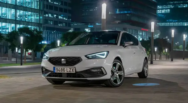 Blanco Seat León aparcado de noche frente a edificios modernos. Las luces LED del coche y las farolas crean un ambiente urbano. Opiniones sobre este modelo son muy positivas.