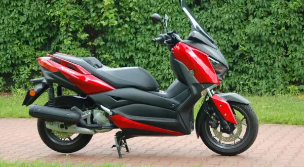 Czerwony skuter Yamaha XMAX 125, gotowy do odblokowania pełnej mocy, zaparkowany na tle zielonego żywopłotu.