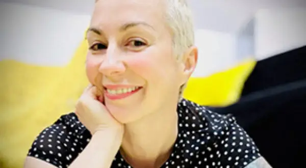 Laura Veltz, a blonde woman with short hair, smiles and rests her chin on her hand. She wears a black polka dot shirt.