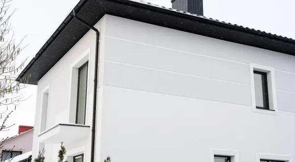 Nowoczesny dom kostka z jasną elewacją, czarnym dachem i kominem. Śnieg na dachu i gałęziach drzew.