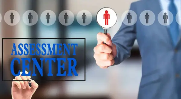 Uomo in giacca blu esamina candidati con lente d'ingrandimento. Testo "ASSESSMENT CENTER" scritto su schermo.