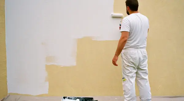 Un peintre en blanc est en train d'enduire sur peinture une partie d'un mur extérieur jaune avec un rouleau blanc.