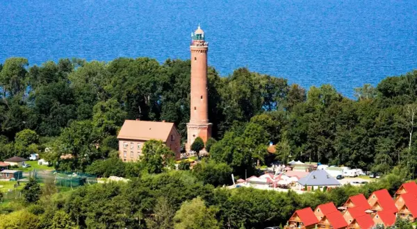 Latarnia morska Sarbinowo góruje nad zielonymi drzewami, z błękitnym morzem w tle. Obok niej budynki i czerwone dachy domków letniskowych.