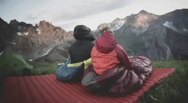 Dos personas sentadas en un colchón inflable, envueltas en sacos de dormir, disfrutan de un paisaje montañoso. El material vivac les permite acampar cómodamente.