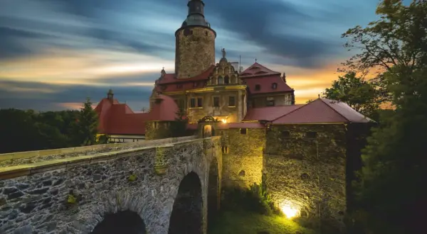 Zamek Czocha, gdzie jest, w blasku zachodzącego słońca i nocnych świateł, z kamiennym mostem i wieżą.