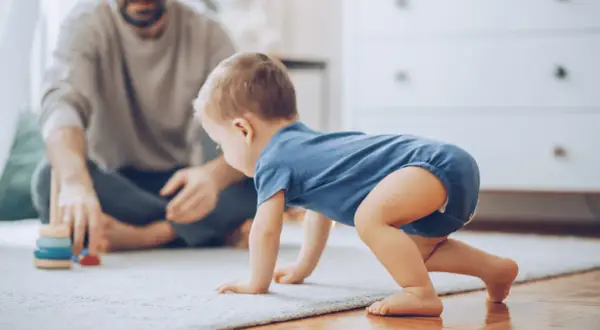 Dziecko w niebieskim body raczkuje na dywanie, obok siedzi ojciec.