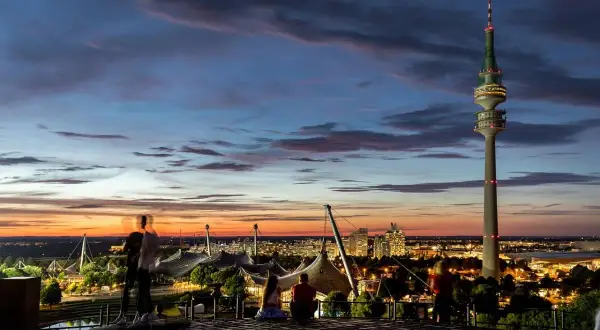 Tramonto a Monaco, Germania. La torre Olympiaturm domina il panorama, con le luci della città che iniziano ad accendersi, un'atmosfera da night club.