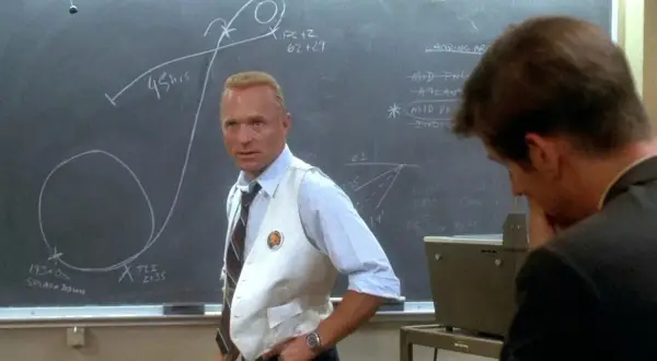 Gene Kranz, w białej kamizelce, omawia trajektorię misji Apollo 13 na tablicy.