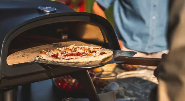 En pizza på muurikka är på väg in i en utomhusugn. Solen skiner och en person i blå skjorta står i bakgrunden.