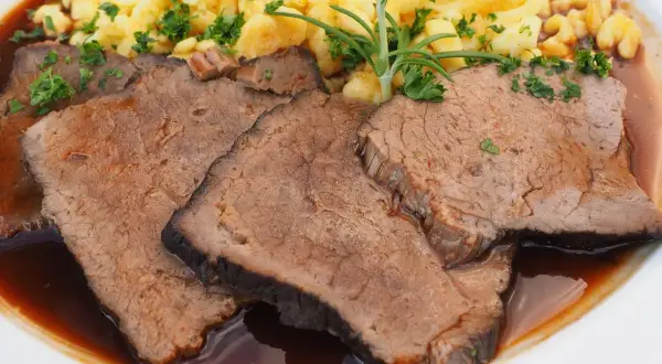 Fertig eingelegter Sauerbraten zubereiten: Zarte Scheiben Rindfleisch in dunkler Sauce mit Spätzle und Rosmarin garniert.