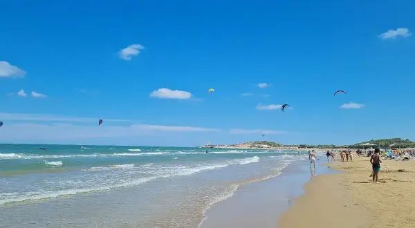 Spiaggia di Scialmarino con kitesurfer che danzano nel cielo azzurro e bagnanti che passeggiano sulla sabbia dorata.