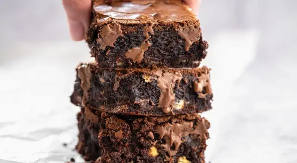 En hand lyfter en stapel saftiga, mörka glutenfria brownies med smält choklad.
