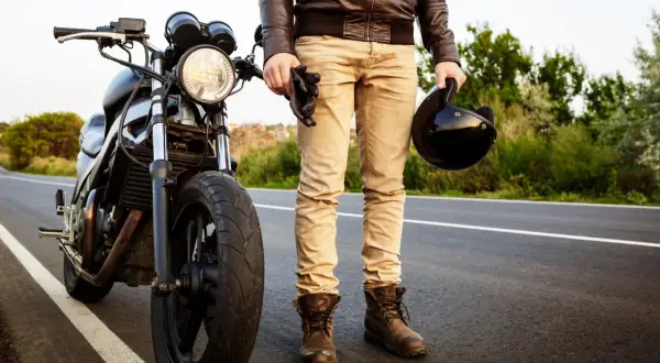 Motocyklista w skórzanej kurtce i beżowych spodniach trzyma kask i rękawice, stojąc obok motocykla na drodze.