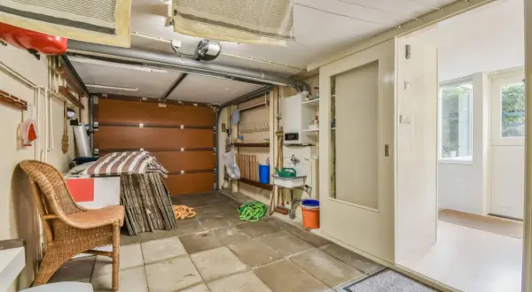 Un garage en cours de transformation en habitation. On y voit une porte de garage, un évier, une chaise en rotin et des éléments de rangement.
