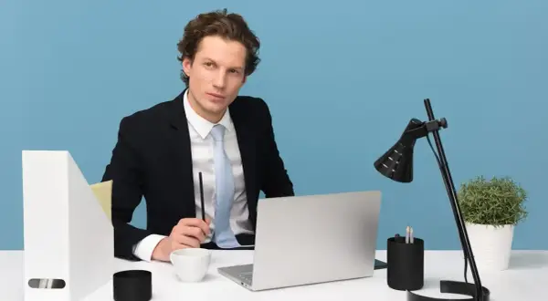 Młody manager w garniturze siedzi przy biurku, trzymając długopis. Na biurku laptop, filiżanka i lampka.