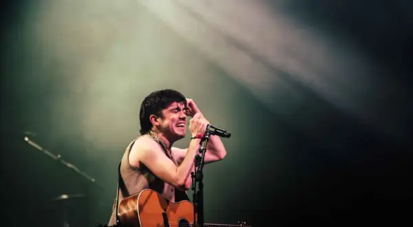 Declan McKenna passionately sings into a microphone, guitar in hand, bathed in stage lights. A glimpse of his setlist is visible.