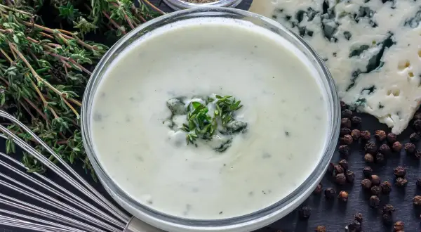 Crema suave y sedosa, lista para tu salsa roquefort receta. Ingredientes frescos como tomillo y pimienta negra completan la escena.
