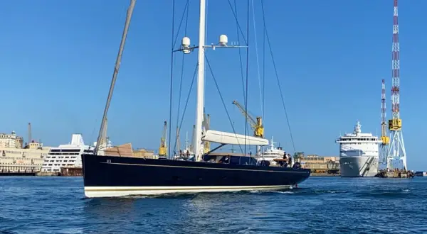 Il kauris iv yacht, scuro e imponente, naviga in un porto con gru e navi da crociera sullo sfondo.