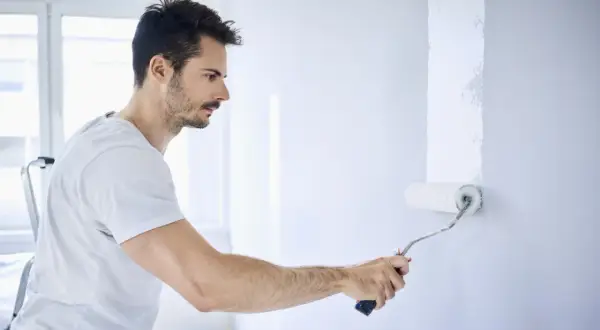 Mann streicht Wand mit weißer Farbe. Die Nachteile von Latexfarbe sind, dass sie schwer zu entfernen ist.