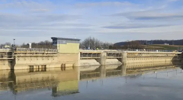 Betonowa zapora w Sieniawie w budowie, z widocznym budynkiem sterowni i rusztowaniami. Woda odbija konstrukcję.
