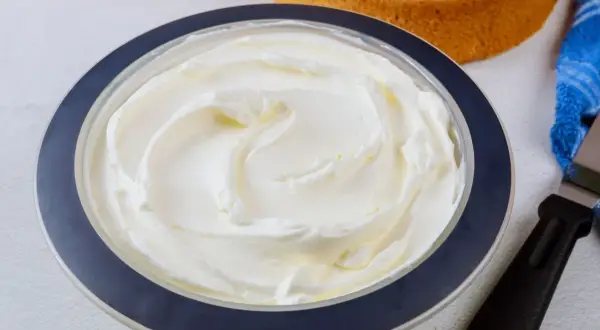Puszysta bita śmietana z mascarpone w misce, gotowa do dekorowania tortu. Obok leżą biszkopty i szpatułka.