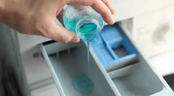 Mano versa liquido blu nel cassetto della lavatrice, mostrando dove mettere il Calgon per un bucato impeccabile.
